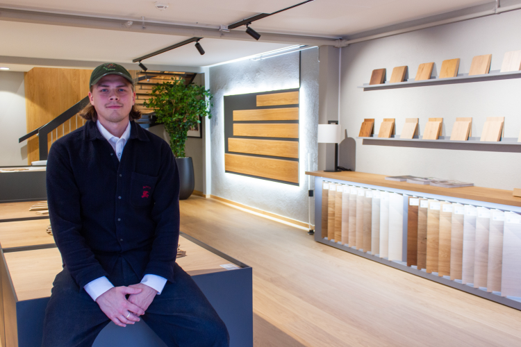 mann i mørke klær og med caps på hodet sitter på benk foran gulvprøver i showrom