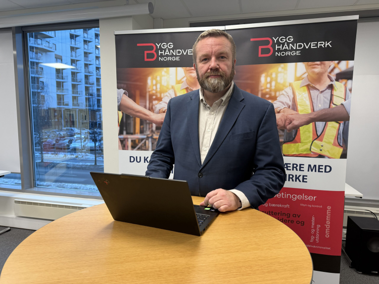 mann med pc på rundt bord foran plakater med reklame for Bygghåndverk Norge