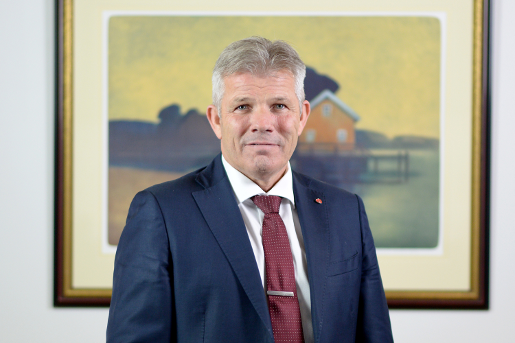 mann i dress og slips foran maleri med gul husgavl