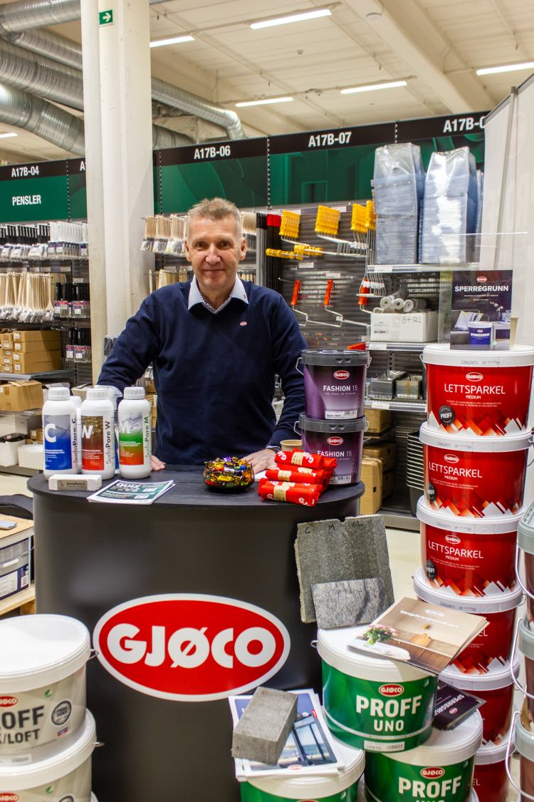 Mann i blå genser bak bord merket Gjøco, viser fram maling og sparkel