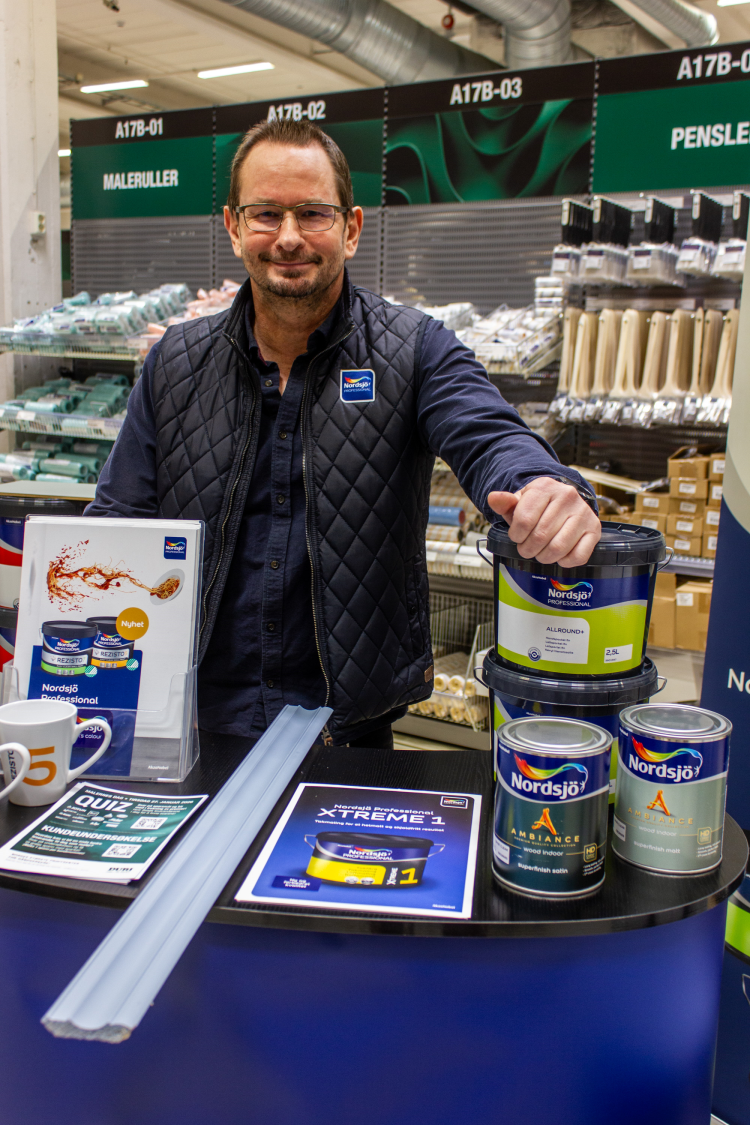 Mann i blå boblejakke og briller bak høyt bord med malt blå list og flere malingspann