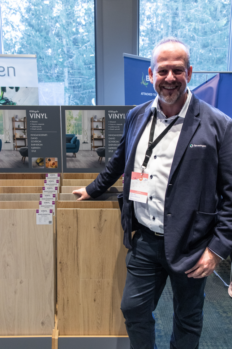 mann i dressjakke poserer ved stativ med gulvprøver