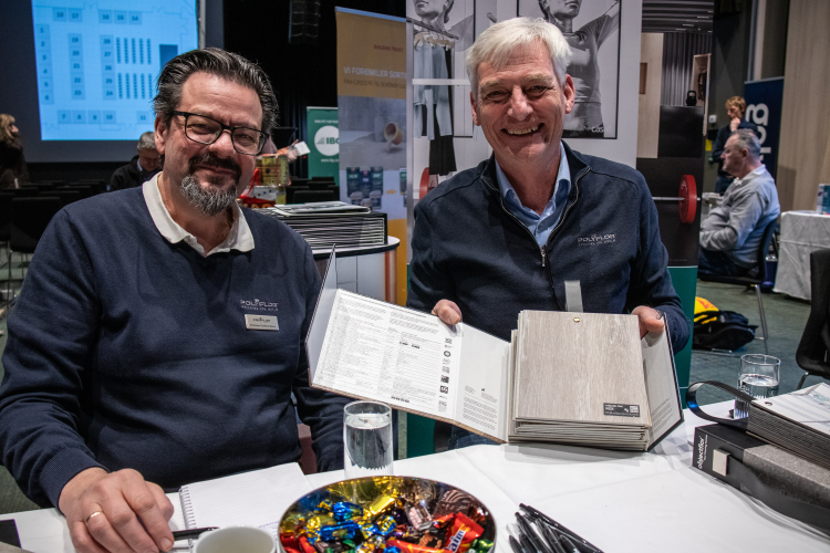 to smilende menn ved et bord, den ene holder en prøvebok med gulvprøver