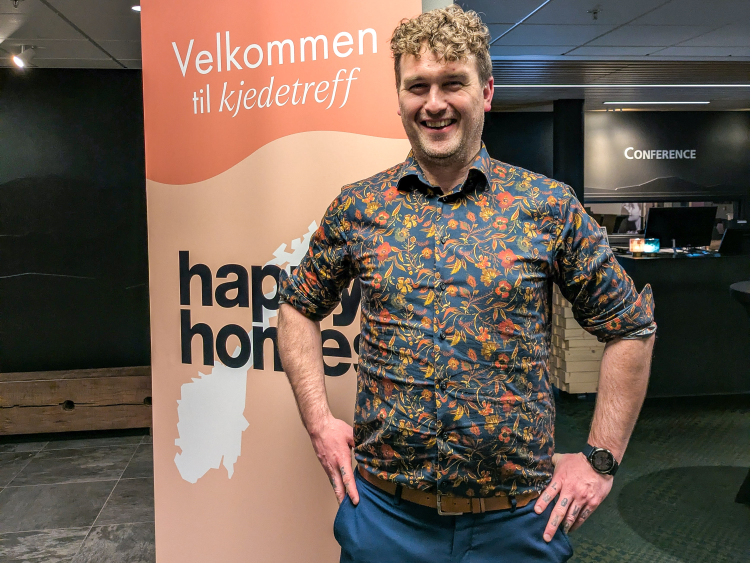 Smilende mann i blomstrete skjorte foran skilt med teksten Happy Homes kjedetreff
