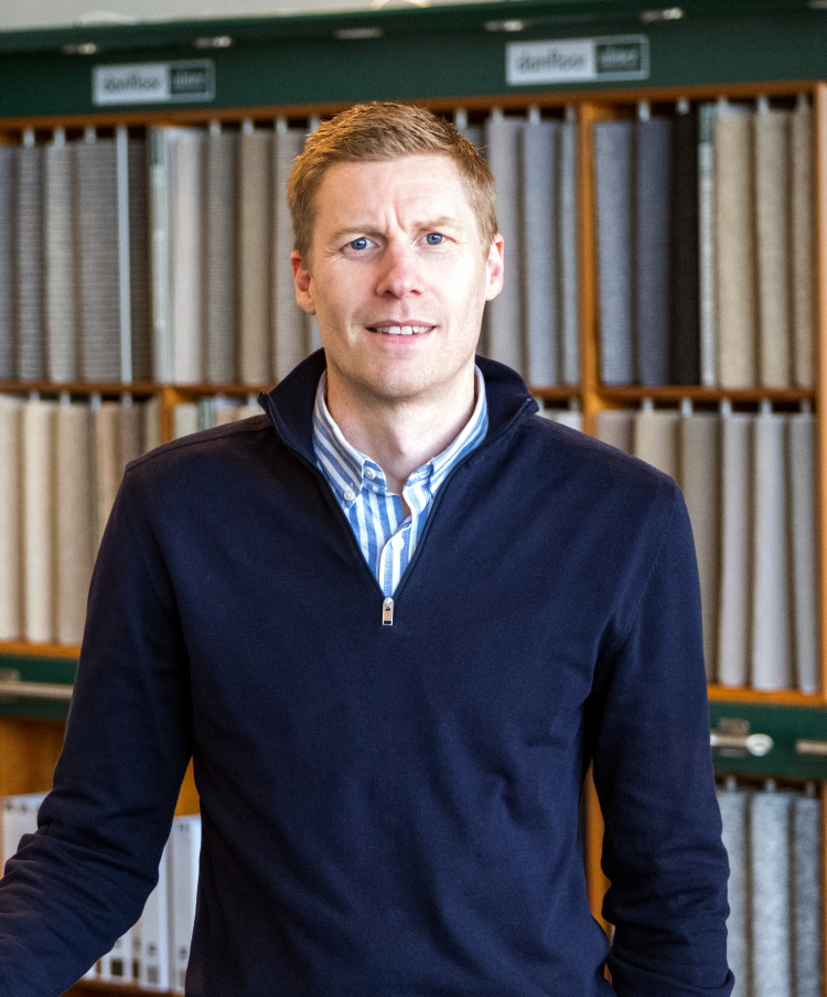 mann i blå genser og stripete skjorte foran teppeprøver