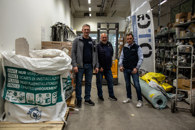 inne på et gulvlager står tre menn ved siden av storsekker med avfall