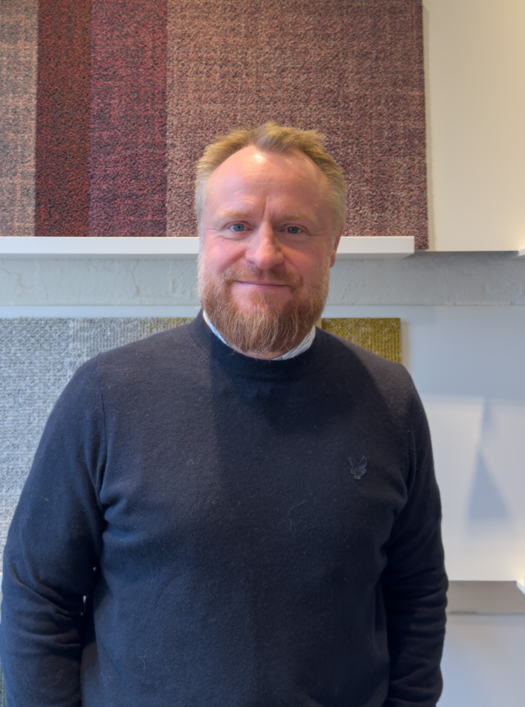 mann med rødlig hår og skjegg kledd i blå genser foran teppeprøver